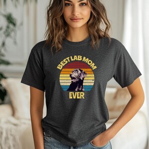 May include: A dark charcoal gray short-sleeve t-shirt featuring a circular graphic with a retro sunset background in orange, yellow, and blue. The graphic displays a black Labrador retriever and the yellow text "BEST LAB MOM EVER".
