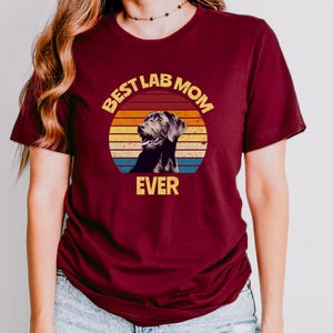 May include: A maroon short-sleeved t-shirt featuring a graphic of a black Labrador retriever inside a circular sunset design with horizontal stripes in yellow, orange, and blue. The text "BEST LAB MOM EVER" is printed in yellow block letters.