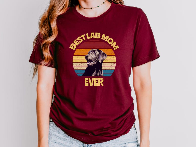 May include: A maroon short-sleeved t-shirt featuring a graphic of a black Labrador retriever inside a circular sunset design with horizontal stripes in yellow, orange, and blue. The text "BEST LAB MOM EVER" is printed in yellow block letters.