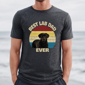 May include: Dark grey t-shirt with a graphic of a black Labrador Retriever and the text "BEST LAB DAD EVER" in a circular design. The design uses a retro colour scheme of red, yellow, and blue.