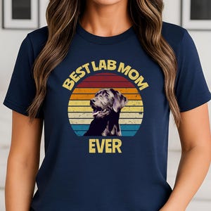 May include: A navy blue short-sleeve t-shirt featuring a graphic of a black Labrador retriever inside a retro-style sunset circle. The text "BEST LAB MOM EVER" is printed in yellow block letters above and below the circular graphic design.