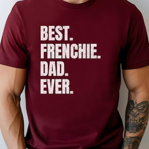 May include: A maroon t-shirt with the text "BEST. FRENCHIE. DAD. EVER." in a bold, white, distressed font. The shirt is short-sleeved and appears to be made of a soft material.