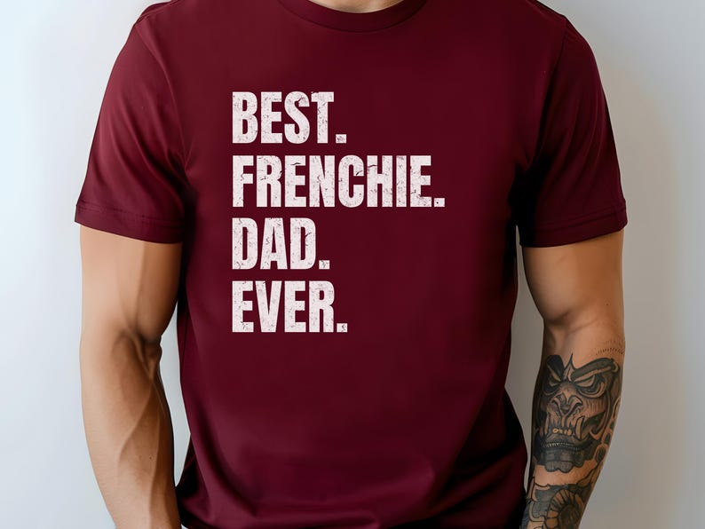 May include: A maroon t-shirt with the text "BEST. FRENCHIE. DAD. EVER." in a bold, white, distressed font. The shirt is short-sleeved and appears to be made of a soft material.