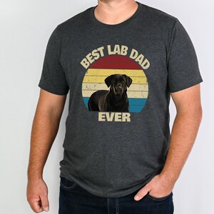 May include: Dark grey t-shirt with a graphic of a black Labrador Retriever inside a retro circle. The circle has a red, yellow, and blue colour scheme, with the text "BEST LAB DAD EVER" in a vintage font.