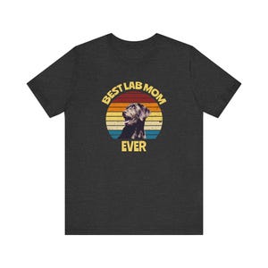 May include: A dark heather gray short-sleeve t-shirt featuring a circular graphic with a sunset background of orange, yellow, and blue stripes. The graphic includes a black Labrador dog illustration and the yellow text BEST LAB MOM EVER.