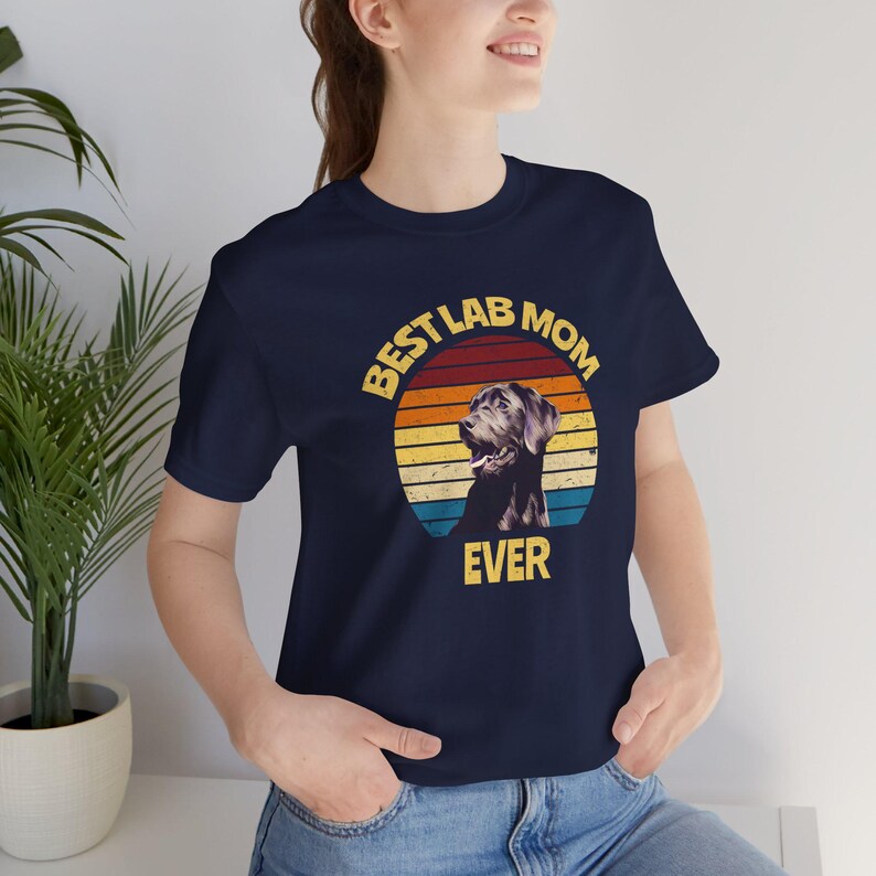 May include: A navy blue short-sleeve t-shirt featuring a graphic of a Labrador retriever inside a sunset circle. The text "BEST LAB MOM EVER" is printed in yellow around the graphic, which includes horizontal stripes of red, orange, and blue.