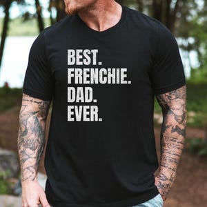 May include: Black t-shirt with the text "BEST. FRENCHIE. DAD. EVER." in white, distressed lettering. The shirt has a v-neck and short sleeves. The person wearing the shirt has tattoos on their arms.