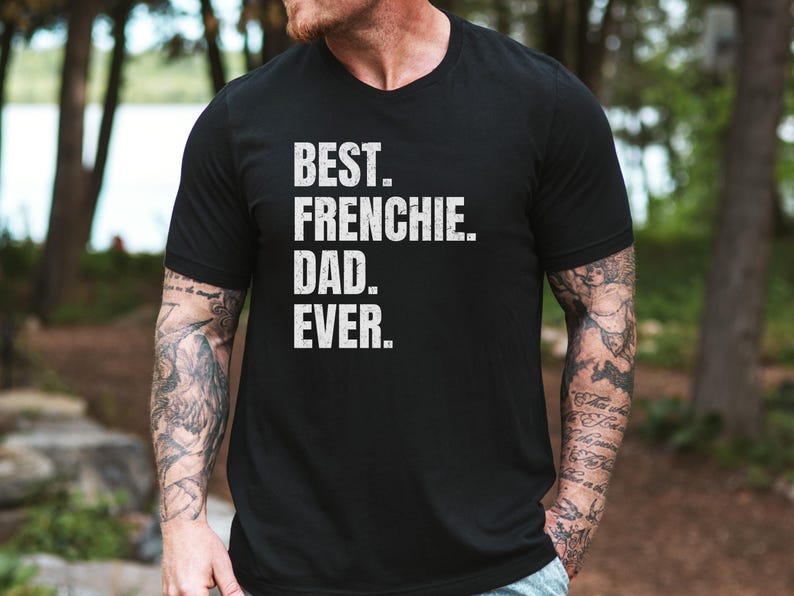 May include: Black t-shirt with the text "BEST. FRENCHIE. DAD. EVER." in white, distressed lettering. The shirt has a v-neck and short sleeves. The person wearing the shirt has tattoos on their arms.