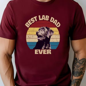 以下が含まれることがあります： レトロな円の中にラブラドールレトリバーのグラフィックが描かれたマルーンのTシャツ。円には赤、黄、青の横縞があり、「BEST LAB DAD EVER」の文字が書かれています。