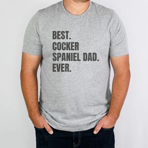 K&ouml;nnte beinhalten: Hellgraues T-Shirt mit dem schwarzen Aufdruck "BEST. COCKER SPANIEL DAD. EVER.". Das Shirt hat einen Rundhalsausschnitt und kurze &Auml;rmel. Die Person, die das Shirt tr&auml;gt, steht vor einem wei&szlig;en Hintergrund.