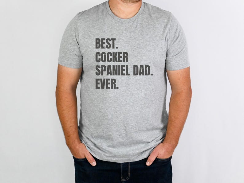 K&ouml;nnte beinhalten: Hellgraues T-Shirt mit dem schwarzen Aufdruck "BEST. COCKER SPANIEL DAD. EVER.". Das Shirt hat einen Rundhalsausschnitt und kurze &Auml;rmel. Die Person, die das Shirt tr&auml;gt, steht vor einem wei&szlig;en Hintergrund.
