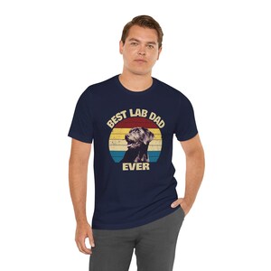 以下が含まれることがあります： ネイビーブルーのTシャツ。ラブラドールレトリバーのグラフィックと「BEST LAB DAD EVER」の文字が円形デザインで描かれています。犬のグラフィックは白黒で、レトロな夕日の背景に配置されています。