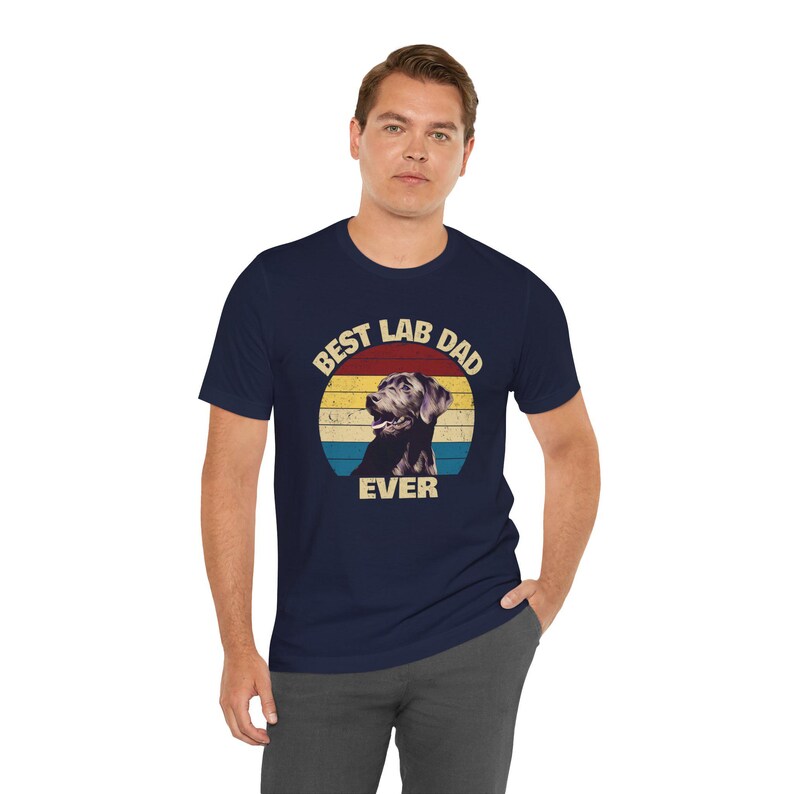 以下が含まれることがあります： ネイビーブルーのTシャツ。ラブラドールレトリバーのグラフィックと「BEST LAB DAD EVER」の文字が円形デザインで描かれています。犬のグラフィックは白黒で、レトロな夕日の背景に配置されています。