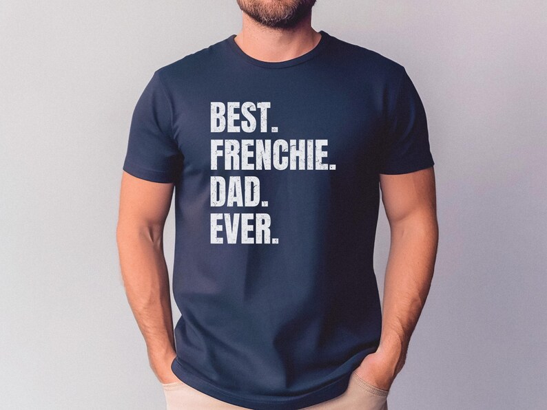 May include: Navy blue t-shirt with the text "BEST. FRENCHIE. DAD. EVER." in white block letters. The shirt is a crew neck and appears to be made of a soft material. The shirt is being worn by a person.