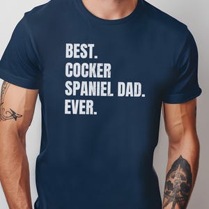 K&ouml;nnte beinhalten: Marineblaues T-Shirt mit dem wei&szlig;en Text "BEST. COCKER SPANIEL DAD. EVER.". Das T-Shirt hat einen Rundhalsausschnitt und wird von einer Person mit T&auml;towierungen an den Armen getragen. Ein l&auml;ssiges Kleidungsst&uuml;ck.
