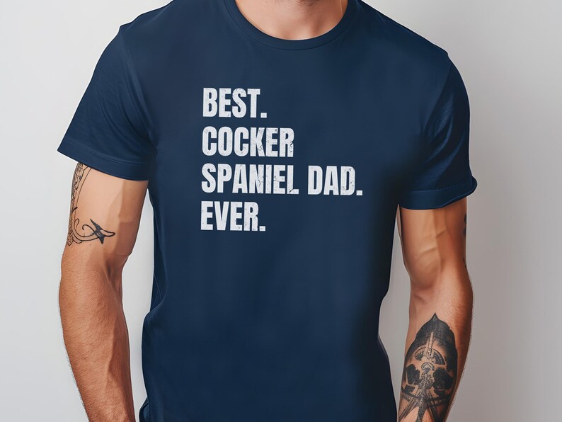 K&ouml;nnte beinhalten: Marineblaues T-Shirt mit dem wei&szlig;en Text "BEST. COCKER SPANIEL DAD. EVER.". Das T-Shirt hat einen Rundhalsausschnitt und wird von einer Person mit T&auml;towierungen an den Armen getragen. Ein l&auml;ssiges Kleidungsst&uuml;ck.