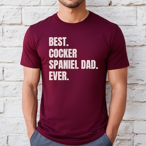 K&ouml;nnte beinhalten: Ein weinrotes T-Shirt mit dem Text "BEST. COCKER SPANIEL DAD. EVER." in wei&szlig;en Blockbuchstaben. Das Shirt ist kurz&auml;rmlig und hat einen Rundhalsausschnitt.