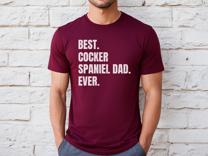 K&ouml;nnte beinhalten: Ein weinrotes T-Shirt mit dem Text "BEST. COCKER SPANIEL DAD. EVER." in wei&szlig;en Blockbuchstaben. Das Shirt ist kurz&auml;rmlig und hat einen Rundhalsausschnitt.