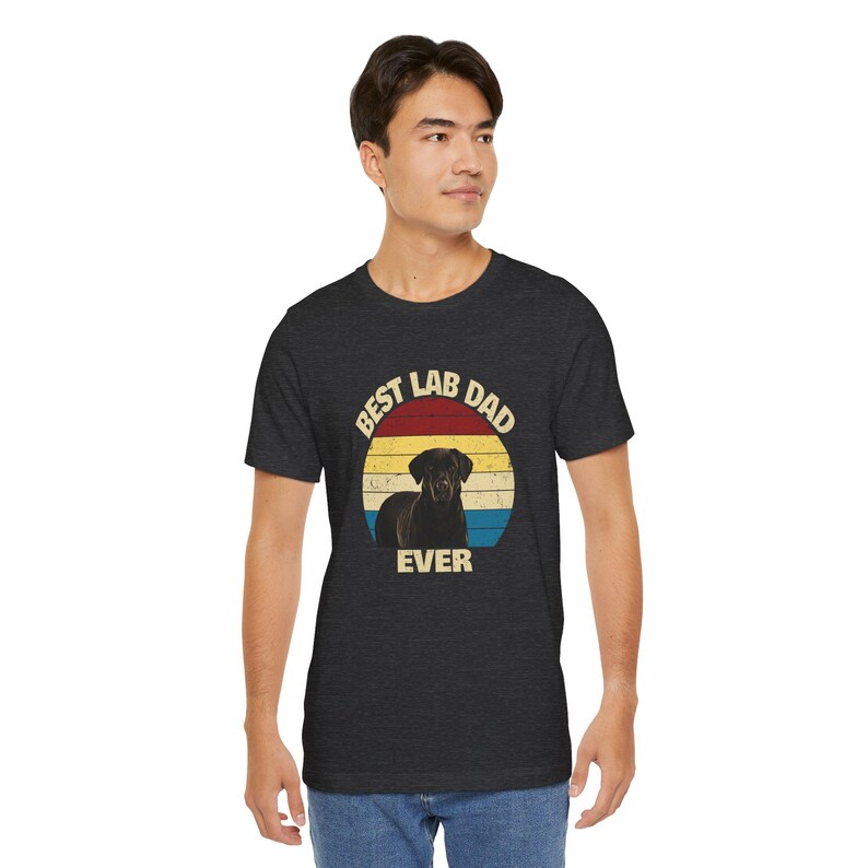 May include: Dark grey t-shirt with a circular graphic. The graphic features a black Labrador Retriever and the text "Best Lab Dad Ever" in a retro style. The graphic has a red, yellow, and blue striped background.