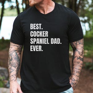 K&ouml;nnte beinhalten: Schwarzes V-Ausschnitt-T-Shirt mit dem wei&szlig;en Text "BEST. COCKER SPANIEL DAD. EVER.". Das Shirt wird von einer Person mit T&auml;towierungen an den Armen getragen. Der Hintergrund ist eine verschwommene Au&szlig;enszene mit B&auml;umen.