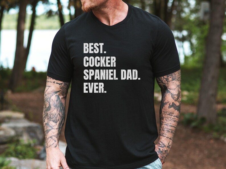 K&ouml;nnte beinhalten: Schwarzes V-Ausschnitt-T-Shirt mit dem wei&szlig;en Text "BEST. COCKER SPANIEL DAD. EVER.". Das Shirt wird von einer Person mit T&auml;towierungen an den Armen getragen. Der Hintergrund ist eine verschwommene Au&szlig;enszene mit B&auml;umen.