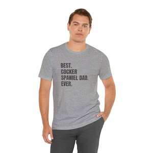 K&ouml;nnte beinhalten: Ein meliertes graues T-Shirt mit dem schwarzen Text "BEST. COCKER SPANIEL DAD. EVER.". Das T-Shirt wird von einer Person getragen.
