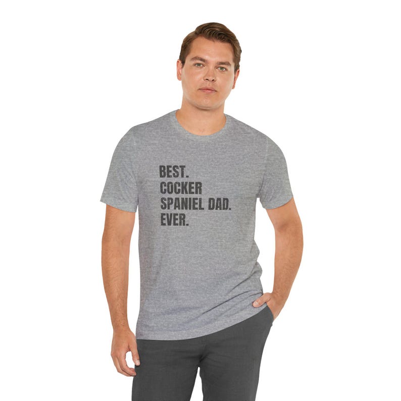 K&ouml;nnte beinhalten: Ein meliertes graues T-Shirt mit dem schwarzen Text "BEST. COCKER SPANIEL DAD. EVER.". Das T-Shirt wird von einer Person getragen.