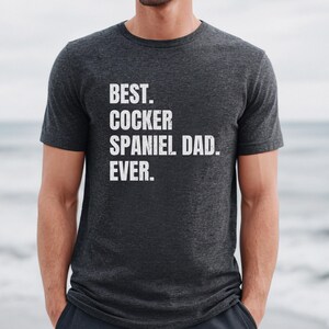 K&ouml;nnte beinhalten: Dunkelgraues T-Shirt mit dem wei&szlig;en Text "BEST. COCKER SPANIEL DAD. EVER.". Das T-Shirt hat einen Rundhalsausschnitt. Die Person, die das T-Shirt tr&auml;gt, steht vor einem verschwommenen Ozeanhintergrund.