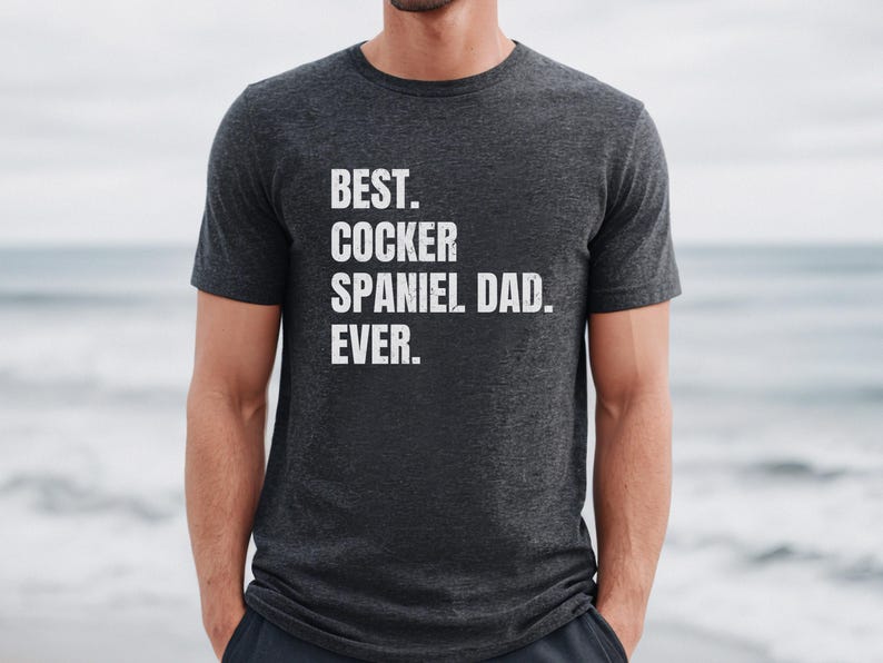 K&ouml;nnte beinhalten: Dunkelgraues T-Shirt mit dem wei&szlig;en Text "BEST. COCKER SPANIEL DAD. EVER.". Das T-Shirt hat einen Rundhalsausschnitt. Die Person, die das T-Shirt tr&auml;gt, steht vor einem verschwommenen Ozeanhintergrund.
