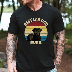 May include: Black t-shirt with a graphic of a black Labrador Retriever in a circular design with red, yellow, and blue stripes. The text "BEST LAB DAD EVER" is arched above and below the dog image.
