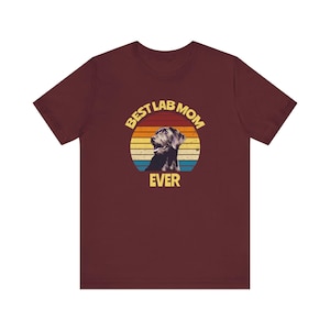 May include: A maroon short-sleeve t-shirt featuring a circular graphic with a sunset-inspired background of yellow, orange, and blue stripes. The graphic includes a black Labrador dog silhouette and the yellow text BEST LAB MOM EVER.
