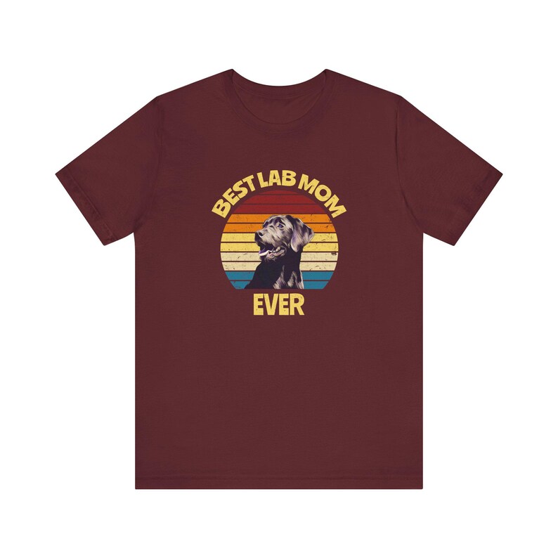 May include: A maroon short-sleeve t-shirt featuring a circular graphic with a sunset-inspired background of yellow, orange, and blue stripes. The graphic includes a black Labrador dog silhouette and the yellow text BEST LAB MOM EVER.