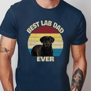 May include: Dark blue t-shirt with a retro design. It features a black Labrador Retriever inside a sunset circle. The text reads "BEST LAB DAD EVER" in a vintage font, ideal for dog lovers.