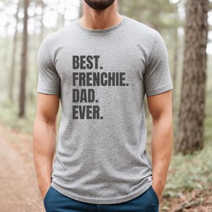 May include: A heather gray t-shirt with the text "BEST. FRENCHIE. DAD. EVER." printed in black. The shirt is short-sleeved and is being worn by a person standing in a forest setting. The person is wearing blue pants.