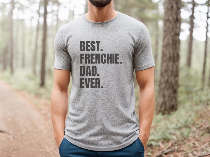 May include: A heather gray t-shirt with the text "BEST. FRENCHIE. DAD. EVER." printed in black. The shirt is short-sleeved and is being worn by a person standing in a forest setting. The person is wearing blue pants.