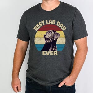 以下が含まれることがあります： レトロなデザインの濃いグレーのTシャツ。「BEST LAB DAD EVER」の文字が、ラブラドールレトリバーのイラストが入った円形デザインで表示されています。デザインには赤、黄、青のストライプが含まれています。Tシャツは人が着ています。