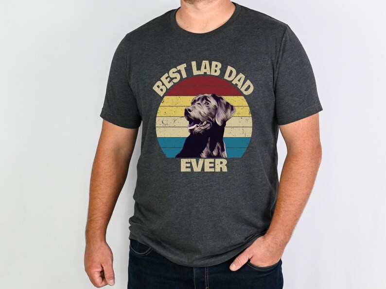 以下が含まれることがあります： レトロなデザインの濃いグレーのTシャツ。「BEST LAB DAD EVER」の文字が、ラブラドールレトリバーのイラストが入った円形デザインで表示されています。デザインには赤、黄、青のストライプが含まれています。Tシャツは人が着ています。