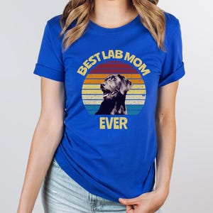May include: A royal blue short-sleeved t-shirt featuring a circular graphic with a retro sunset background in orange, yellow, and teal. The graphic depicts a black Labrador retriever and includes the yellow text BEST LAB MOM EVER.