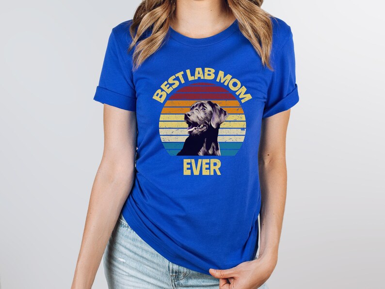 May include: A royal blue short-sleeved t-shirt featuring a circular graphic with a retro sunset background in orange, yellow, and teal. The graphic depicts a black Labrador retriever and includes the yellow text BEST LAB MOM EVER.