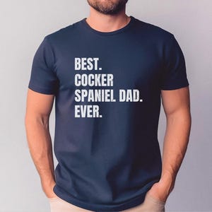 K&ouml;nnte beinhalten: Marineblaues T-Shirt mit dem wei&szlig;en Text "BEST. COCKER SPANIEL DAD. EVER.". Das T-Shirt hat einen Rundhalsausschnitt und wird von einer Person getragen.