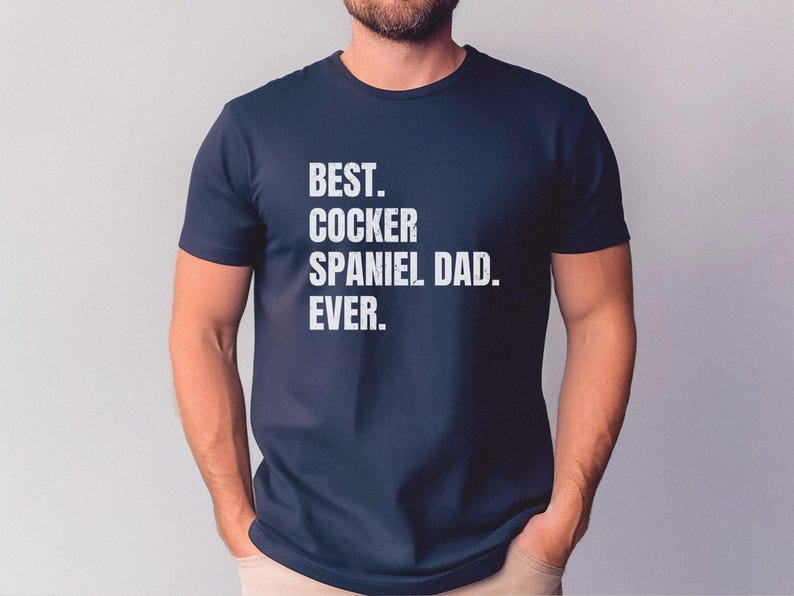 K&ouml;nnte beinhalten: Marineblaues T-Shirt mit dem wei&szlig;en Text "BEST. COCKER SPANIEL DAD. EVER.". Das T-Shirt hat einen Rundhalsausschnitt und wird von einer Person getragen.