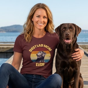 May include: A maroon short-sleeved t-shirt featuring a graphic print of a chocolate Labrador retriever inside a sunset-colored circle. The text BEST LAB MOM EVER is printed in yellow around the graphic. The shirt is paired with blue jeans.