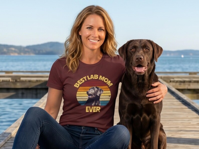 May include: A maroon short-sleeved t-shirt featuring a graphic print of a chocolate Labrador retriever inside a sunset-colored circle. The text BEST LAB MOM EVER is printed in yellow around the graphic. The shirt is paired with blue jeans.