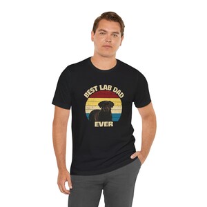May include: Black t-shirt with a retro design featuring a Labrador Retriever and the text "Best Lab Dad Ever" in a circular design with red, yellow, and blue stripes. The shirt is worn by a person.