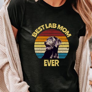 May include: A black t-shirt featuring a graphic of a Labrador retriever inside a circular sunset design with horizontal stripes in yellow, orange, and blue. The text "BEST LAB MOM EVER" is printed in yellow block letters around the graphic.
