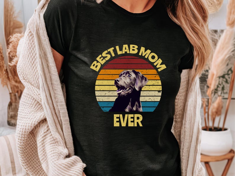 May include: A black t-shirt featuring a graphic of a Labrador retriever inside a circular sunset design with horizontal stripes in yellow, orange, and blue. The text "BEST LAB MOM EVER" is printed in yellow block letters around the graphic.