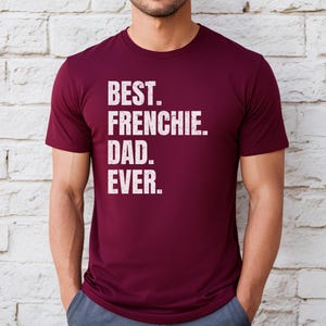 May include: A maroon t-shirt with the text "BEST. FRENCHIE. DAD. EVER." in white, distressed lettering. The shirt is short-sleeved and appears to be made of a soft material. The person is wearing blue pants.