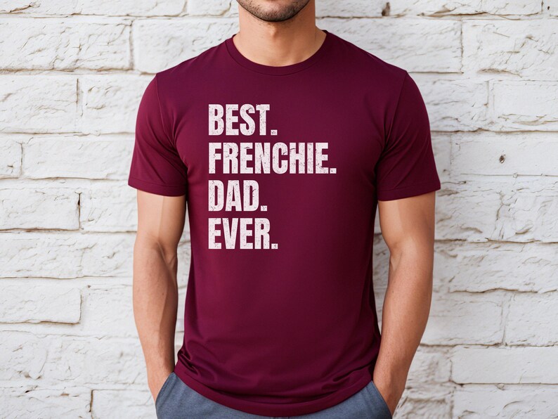 May include: A maroon t-shirt with the text "BEST. FRENCHIE. DAD. EVER." in white, distressed lettering. The shirt is short-sleeved and appears to be made of a soft material. The person is wearing blue pants.