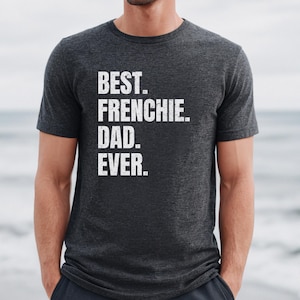 May include: Dark gray t-shirt with the text "BEST. FRENCHIE. DAD. EVER." in white block letters. The shirt is a crew neck style and is being worn by a person. The background is a blurred view of the ocean.