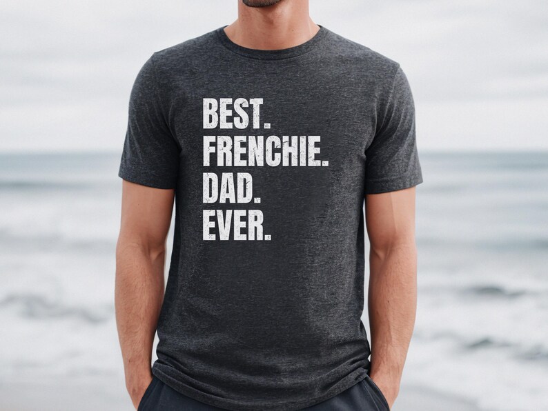May include: Dark gray t-shirt with the text "BEST. FRENCHIE. DAD. EVER." in white block letters. The shirt is a crew neck style and is being worn by a person. The background is a blurred view of the ocean.
